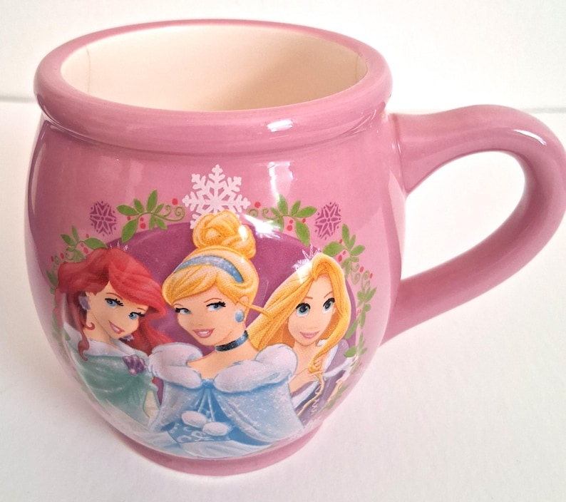 Puede incluir: Taza de cer&aacute;mica rosa con tres princesas de dibujos animados. La taza tiene forma redondeada con asa e interior rosa claro. Las princesas est&aacute;n representadas en un dise&ntilde;o colorido con tem&aacute;tica invernal.
