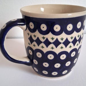 Puede incluir: Una taza de cerámica con exterior azul oscuro, adornada con patrones geométricos blancos y verdes. El asa y el borde también son azul oscuro. El interior es de color crema. La taza está diseñada para bebidas calientes.