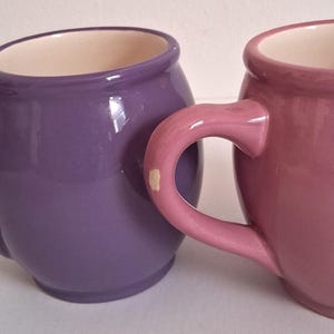 Puede incluir: Dos tazas de cer&aacute;mica, una morada y otra malva, con cuerpos redondeados y asas grandes y curvas. El interior de cada taza es de color crema. Las tazas son brillantes y parecen hechas a mano.
