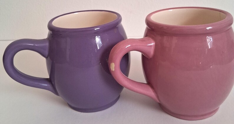 Puede incluir: Dos tazas de cer&aacute;mica, una morada y otra malva, con cuerpos redondeados y asas grandes y curvas. El interior de cada taza es de color crema. Las tazas son brillantes y parecen hechas a mano.