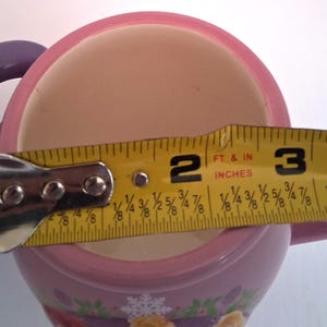Puede incluir: Una taza de cer&aacute;mica rosa y blanca con una cinta m&eacute;trica amarilla que muestra un di&aacute;metro de aproximadamente 7,6 cm. La taza tiene un borde rosa y un dise&ntilde;o decorativo en la base.