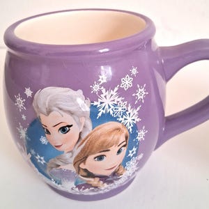 Puede incluir: Una taza de cer&aacute;mica morada con Elsa y Anna de Frozen. La taza tiene forma redondeada con un asa grande y est&aacute; decorada con copos de nieve. El interior es de color crema.