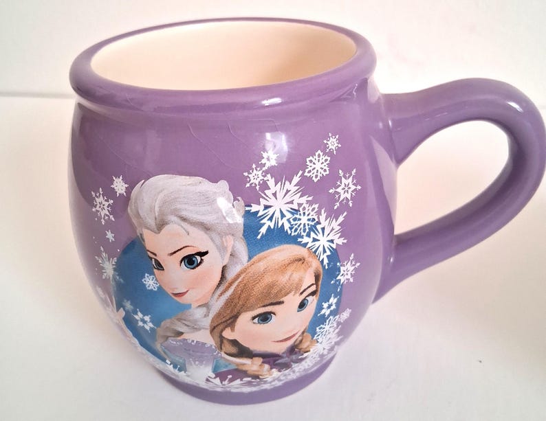 Puede incluir: Una taza de cer&aacute;mica morada con Elsa y Anna de Frozen. La taza tiene forma redondeada con un asa grande y est&aacute; decorada con copos de nieve. El interior es de color crema.