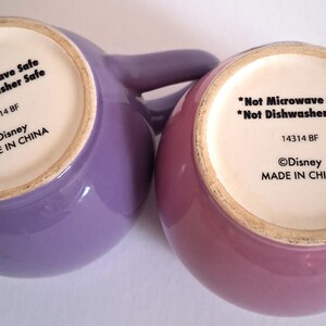 Puede incluir: Se muestran dos tazas de cer&aacute;mica, una lavanda y otra rosa, desde la parte inferior. El texto en la parte inferior de cada taza dice "*Not Microwave Safe *Not Dishwasher Safe 14314 BF &copy;Disney MADE IN CHINA."