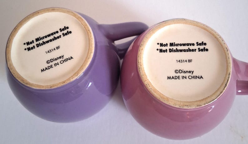 Puede incluir: Se muestran dos tazas de cer&aacute;mica, una lavanda y otra rosa, desde la parte inferior. El texto en la parte inferior de cada taza dice "*Not Microwave Safe *Not Dishwasher Safe 14314 BF &copy;Disney MADE IN CHINA."
