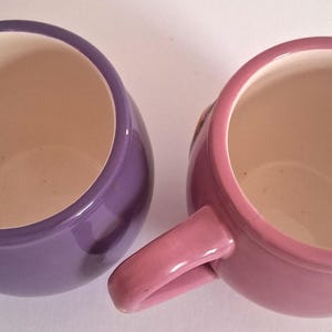 Puede incluir: Vista superior de dos tazas de cer&aacute;mica, una morada y otra rosa. Las tazas tienen forma redondeada con un asa lateral. El interior de cada taza es de color beige claro.