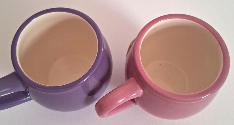 Puede incluir: Vista superior de dos tazas de cer&aacute;mica, una morada y otra rosa. Las tazas tienen forma redondeada con un asa lateral. El interior de cada taza es de color beige claro.