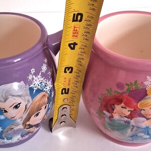 Puede incluir: Dos tazas de cer&aacute;mica, una morada y otra rosa, cada una con interior blanco y asa. La taza morada presenta personajes de Frozen, mientras que la rosa muestra princesas Disney. Las tazas miden unos 10 cm de alto.