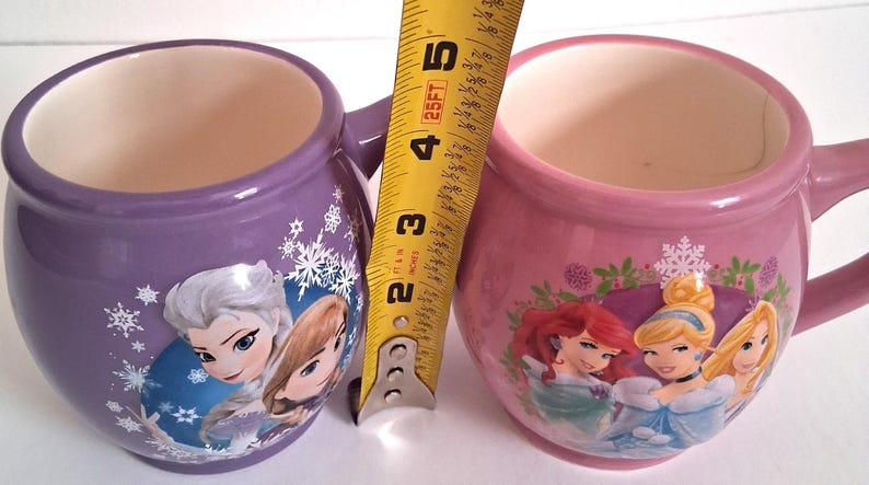 Puede incluir: Dos tazas de cer&aacute;mica, una morada y otra rosa, cada una con interior blanco y asa. La taza morada presenta personajes de Frozen, mientras que la rosa muestra princesas Disney. Las tazas miden unos 10 cm de alto.