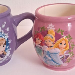 Puede incluir: Dos tazas de cer&aacute;mica, una morada y otra rosa, cada una con forma redondeada y un asa. La taza morada presenta personajes de Frozen, mientras que la taza rosa muestra princesas de Disney. Ambas tazas tienen el interior blanco.