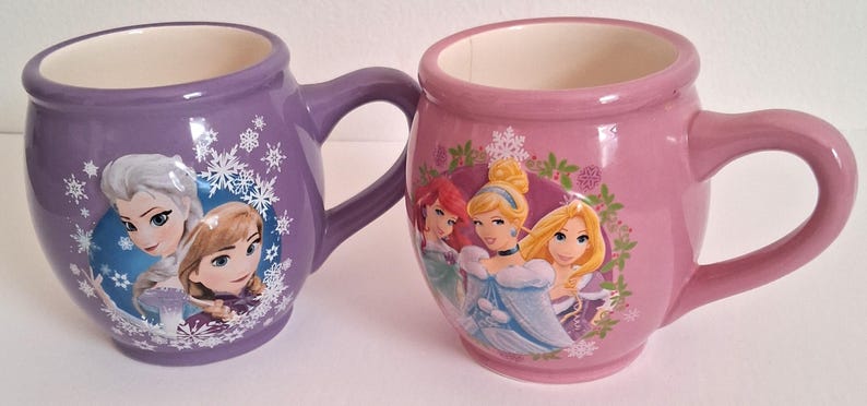 Puede incluir: Dos tazas de cer&aacute;mica, una morada y otra rosa, cada una con forma redondeada y un asa. La taza morada presenta personajes de Frozen, mientras que la taza rosa muestra princesas de Disney. Ambas tazas tienen el interior blanco.