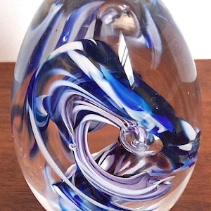 Pode incluir: Um peso de papel de vidro transparente com formato oval. Redemoinhos de vidro azul, branco e roxo estão encapsulados no interior, criando um design abstrato dinâmico. O peso de papel está sobre uma superfície de madeira.