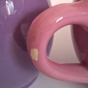 Puede incluir: Primer plano de dos tazas de cer&aacute;mica. Una taza es de color lavanda s&oacute;lido y la otra rosa claro. El asa de la taza rosa est&aacute; enfocada, mostrando una astilla en la pintura. Las tazas parecen ser parte de un juego.