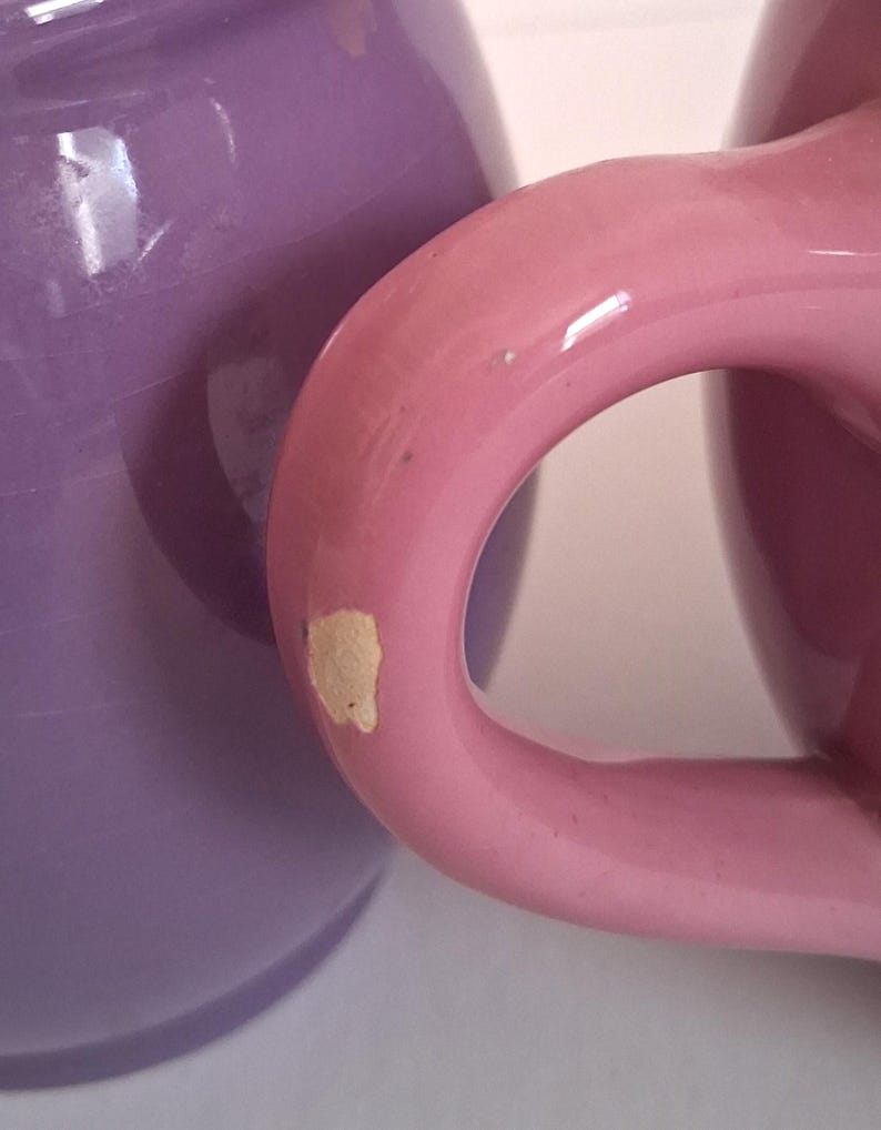Puede incluir: Primer plano de dos tazas de cer&aacute;mica. Una taza es de color lavanda s&oacute;lido y la otra rosa claro. El asa de la taza rosa est&aacute; enfocada, mostrando una astilla en la pintura. Las tazas parecen ser parte de un juego.