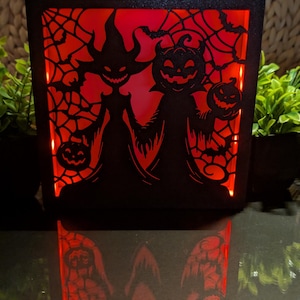 Könnte beinhalten: Eine rechteckige schwarze Leuchtbox mit einer lasergeschnittenen Silhouette einer Hexe und einer Figur mit Kürbiskopf, die Kürbislaternen vor einem leuchtend roten Hintergrund hält. Das komplexe Design zeigt Spinnennetze und Fledermäuse.