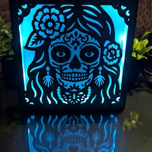 Puede incluir: Una caja de luz cuadrada negra con un diseño cortado a láser de una calavera de azúcar con accesorios florales en el cabello y pendientes colgantes. El diseño está retroiluminado con una luz azul brillante de alto contraste.