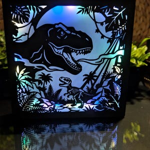 May include: A square black shadow box light featuring a layered paper cutout design of a large T-Rex head and a smaller dinosaur skeleton silhouette set against a glowing blue and purple LED background with palm trees and tropical foliage.