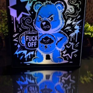 May include: A square light box featuring a graphic of a grumpy blue bear holding a steaming mug with the text FUCK OFF. The bear has a storm cloud symbol on its belly. The design includes black and blue accents with star and splash motifs.
