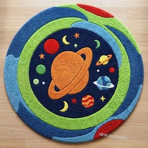 Può includere: Un tappeto rotondo trapuntato con un centro blu scuro raffigurante un grande pianeta arancione con anelli, pianeti più piccoli, stelle e lune crescenti. Il bordo è composto da anelli concentrici in verde brillante, azzurro e rosso. Testo: NEW AIS CARPET.