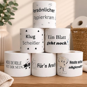Puede incluir: Una pirámide de seis rollos de papel higiénico blancos con texto e ilustraciones impresas en negro. Las frases incluyen Persönlicher Papierkram, Klug Scheißer, Ein Blatt geht noch, Möge die Rolle mit dir sein, Für'n Arsch y Heute schon abgerollt.