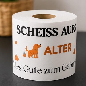 Puede incluir: Un rollo de papel higi&eacute;nico blanco con texto en negro y naranja que dice "SCHEISS AUFS ALTER" y "Alles Gute zum Geburtstag". El dise&ntilde;o incluye una peque&ntilde;a ilustraci&oacute;n de un perro naranja y varios iconos de excrementos naranjas impresos.
