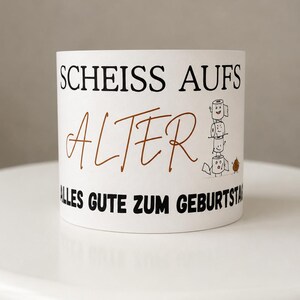 Puede incluir: Una taza de cer&aacute;mica blanca con el texto negro "SCHEISS AUFS" sobre la palabra "ALTER" escrita en letra cursiva marr&oacute;n y el texto negro "ALLES GUTE ZUM GEBURTSTAG". Un gr&aacute;fico de tres rollos de papel higi&eacute;nico apilados con caras.