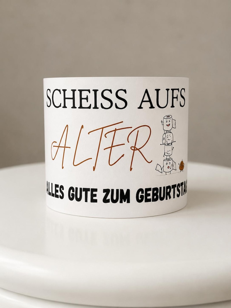 Puede incluir: Una taza de cer&aacute;mica blanca con el texto negro "SCHEISS AUFS" sobre la palabra "ALTER" escrita en letra cursiva marr&oacute;n y el texto negro "ALLES GUTE ZUM GEBURTSTAG". Un gr&aacute;fico de tres rollos de papel higi&eacute;nico apilados con caras.