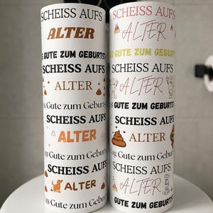 Puede incluir: Dos objetos cil&iacute;ndricos blancos con texto repetido en negro y marr&oacute;n que dice "SCHEISS AUFS ALTER" y "GUTE ZUM GEBURTSTAG". Los dise&ntilde;os incluyen peque&ntilde;as ilustraciones coloridas de personajes de caca, sombreros de fiesta y un perro marr&oacute;n.