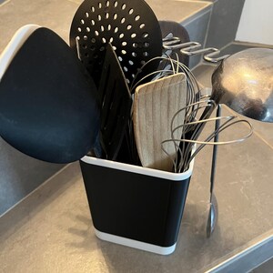 Peut inclure: Un porte-ustensiles de cuisine rectangulaire noir et blanc contenant divers outils de cuisson, dont une spatule ajourée noire, une écumoire noire, une spatule en bois, un fouet en métal et une louche en acier inoxydable à long manche.