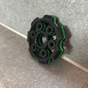 Peut inclure: Un assemblage d'engrenages mécaniques circulaire composé d'un grand anneau extérieur noir avec des dents intégrées et une couche intérieure vert vif. L'objet contient plusieurs petits engrenages noirs imbriqués autour d'une ouverture centrale.