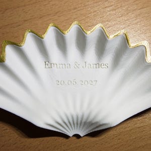 Peut inclure: Un plat blanc en forme de coquillage avec un bord festonné peint en or. Le centre du plat est gravé avec le texte Emma & James et la date 20.06.2027. La surface du plat présente une texture subtile et ondulée rayonnant depuis la base.