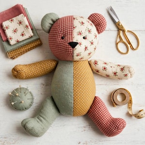 May include: A patchwork teddy bear constructed from various fabrics including red gingham, cream floral print, mustard yellow polka dot, and sage green linen. The bear is surrounded by a stack of fabric squares, metal scissors, a pin cushion, and a tape measure.