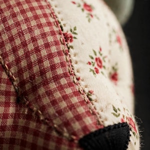 May include: A close-up view of a fabric object featuring a combination of red and white gingham check pattern and a cream-colored fabric with a small red floral print. The two distinct fabric sections are joined by visible tan stitching.