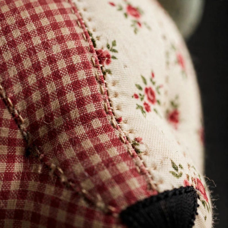 May include: A close-up view of a fabric object featuring a combination of red and white gingham check pattern and a cream-colored fabric with a small red floral print. The two distinct fabric sections are joined by visible tan stitching.