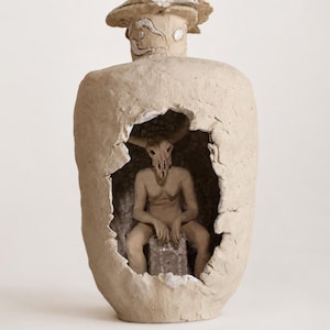 May include: A beige ceramic vessel with a rough, textured surface and a wide, jagged opening in the center. Inside the hollow space sits a sculpted figure with a horned animal skull head, seated on a small block, all in a matching matte beige tone.
