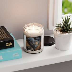 Pode incluir: Uma vela num frasco de vidro transparente com cera branca e um pavio. O rótulo apresenta um design de coração em ganga azul com costuras visíveis e uma pequena etiqueta retangular. A vela está ao lado de um livro preto e uma suculenta verde.