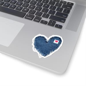 May include: A heart-shaped sticker featuring a dark blue denim fabric texture with frayed, distressed edges and white stitching. A small white rectangular patch with a tiny red heart icon is attached to the upper right side of the denim heart.