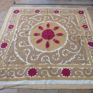 May include: A square textile wall hanging featuring a central magenta floral medallion surrounded by tan scrollwork patterns. The border displays a repeating magenta flower motif set against a tan background, framed by a white outer edge.