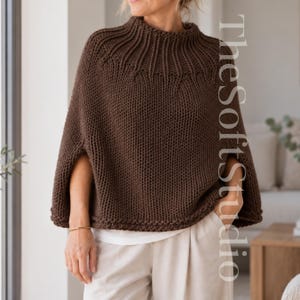 May include: A dark brown knitted poncho featuring a ribbed mock neck collar and a textured garter stitch body. The garment has wide arm openings and is worn over white trousers. The text TheSoftStudio is visible on the right side of the image.