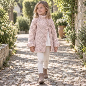 Könnte beinhalten: Eine grob gestrickte Strickjacke in zartem Altrosa mit hohem Kragen, langen Ärmeln mit Umschlagbündchen und einem seitlichen Verschluss mit drei großen, runden, passenden Knöpfen. Das Kleidungsstück wird über einem schlichten weißen Oberteil und einer hellen Hose getragen.