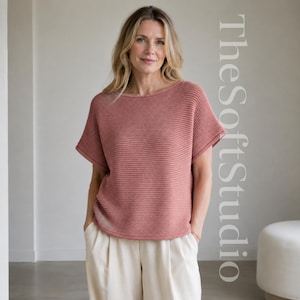 May include: A dusty rose short-sleeved knit jumper with a wide boat neckline and a horizontal ribbed texture. The garment features a relaxed fit with dropped shoulders and a straight hemline. The text TheSoftStudio is visible on the right side.