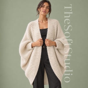 May include: An open-front, cream-coloured knit cardigan with a textured, ribbed pattern and wide, batwing-style sleeves. The garment is worn over a black top and black trousers. The text TheSoftStudio is visible on the right side of the image.