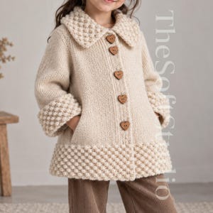 Può includere: Un cappotto in maglia beige con un motivo testurizzato a punto nocciolina su colletto, polsini e orlo. Il capo presenta maniche lunghe, due tasche laterali e cinque bottoni in legno a forma di cuore sul davanti. Il testo TheSoftStudio è visibile.