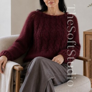 May include: A deep burgundy long-sleeved sweater featuring a textured cable knit pattern on the front, a round neckline, and ribbed cuffs. The sweater has a fuzzy, soft appearance and is paired with grey trousers. The text TheSoftStudio is visible.