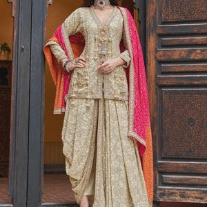 May include: A beige long-sleeved tunic and dhoti-style pants set featuring intricate gold embroidery, beadwork, and hanging tassels. The outfit is paired with a vibrant pink and orange gradient dupatta with a dotted pattern.