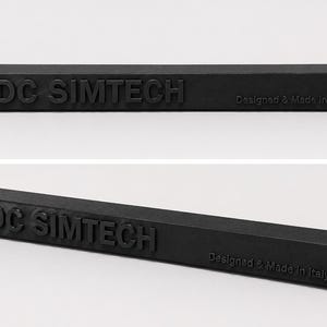 Op de afbeelding: Een matzwarte rechthoekige balk met in reliëf aangebrachte tekst DC SIMTECH en Designed & Made in Italy. De balk heeft aan beide uiteinden aan de bovenzijde een gat. Het object wordt getoond vanuit een vooraanzicht en een schuin perspectief.