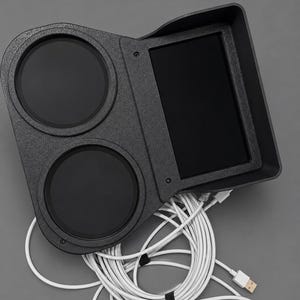 Op de afbeelding: Een zwarte, getextureerde consolehouder met twee ronde bekerhouders en een rechthoekige, verzonken schermbehuizing. Meerdere witte USB-kabels zijn gebundeld en bevestigd aan de onderkant van de unit, met één zichtbare USB-A-connector.