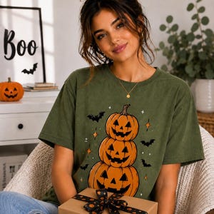 Puede incluir: Una camiseta de manga corta color verde oliva con un estampado gráfico de tres calabazas de Halloween naranjas apiladas con caras negras talladas, rodeadas de pequeños murciélagos negros y estrellas de cuatro puntas en el frente.