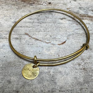 Op de afbeelding: Een goudkleurige metalen armband met een dunne, uitrekbare draadband en een enkele, ronde, platte bedel die aan een klein lusje hangt. De bedel is gestempeld met de tekst SLAVETINA in hoofdletters en heeft een textuurrijke, geborstelde afwerking.