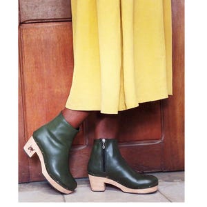 May include: Olive green leather ankle boots with a wooden block heel and sole. The boots feature a side zipper closure, a rounded toe, and a row of small metal studs securing the leather upper to the light-colored wooden base of the footwear.